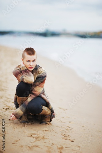 Young beautiful girl with very short hair in a knitted cardigan sits on his haunches on the sandy Bank of the Neva river autumn on a chilly day.
