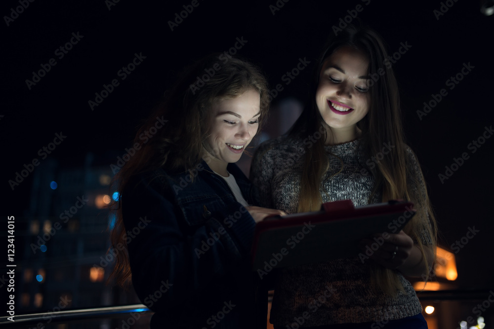 Two women look down to digital tablet in balcony at night Two women look down to digital tablet in balcony at night