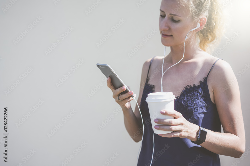 Sunny summer day,young woman dressed in blue silk top,standing near a white wall and listening to music on headphones.In one hand she holds smartphone in other-cup with a drink.Girl using gadget. Sunny summer day,young woman dressed in blue silk top,standing near a white wall and listening to music on headphones.In one hand she holds smartphone in other-cup with a drink.Girl using gadget.