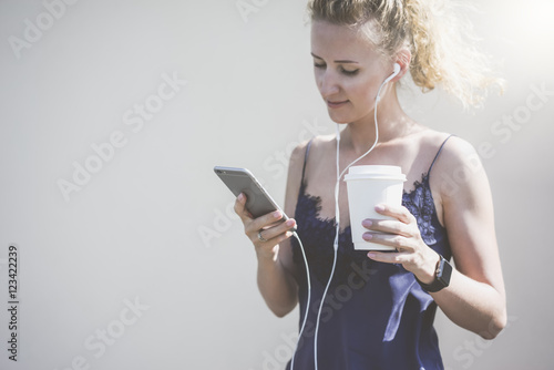 Girl with blond hair,wearing blue top,standing and using smartphone while listening to music on headphones.In her hand she holds paper cup with drink.In the background a white wall.Girl using gadget.