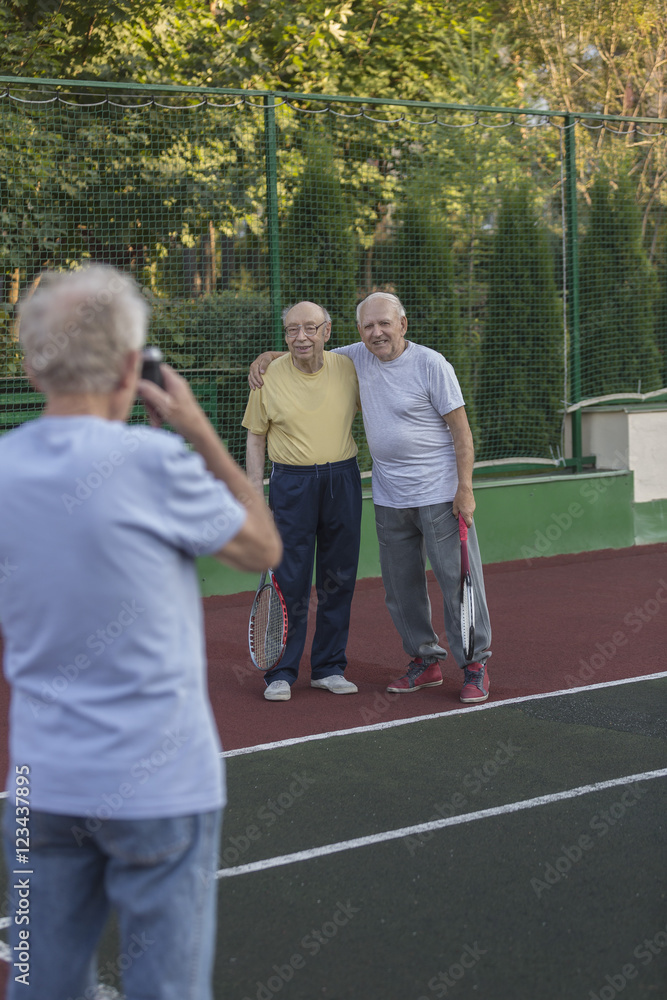 Rear view of man photographing friends standing against fence at tennis court Rear view of man photographing friends standing against fence at tennis court