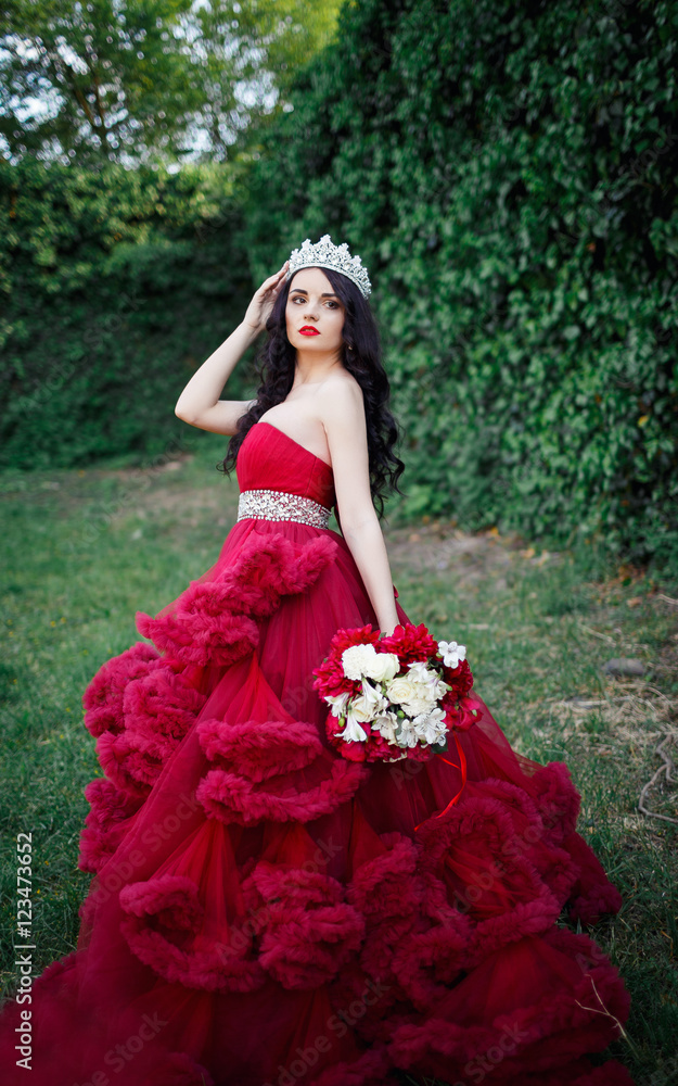 beautiful brunette in a crown with a bouquet in his hand and in a chic big and lush red dress in the park among the green beautiful brunette in a crown with a bouquet in his hand and in a chic big and lush red dress in the park among the green