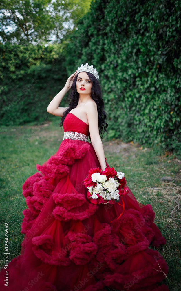 beautiful brunette in a crown with a bouquet in his hand and in a chic big and lush red dress in the park among the green