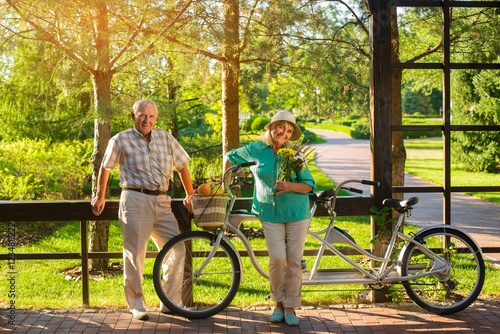 Senior couple near tandem bicycle. Woman with bouquet smiling. Anniversary of wedding. Save warm feelings.