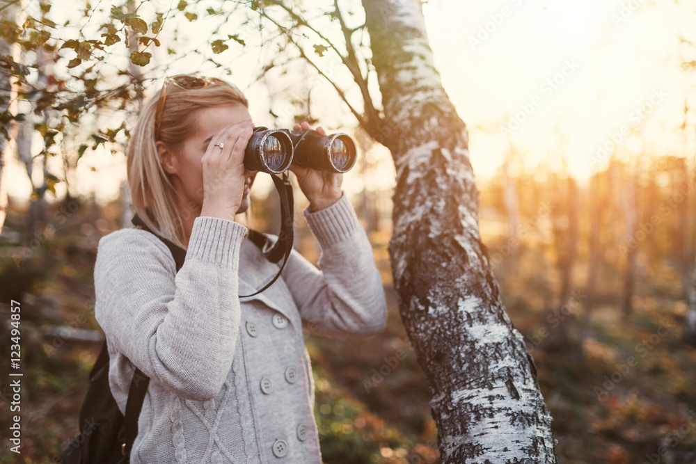 Naklejka premium Beautiful happy blonde woman enjoying autumn outdoors. She holding binoculars and looking at gorgeous landscape and sunset.