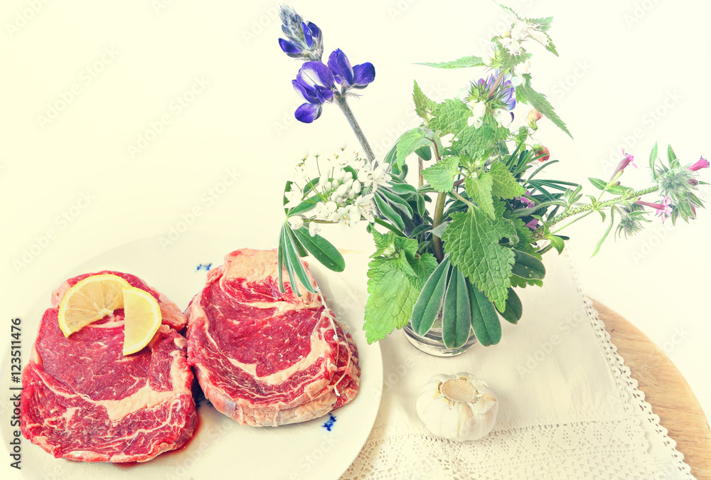 Raw beef steak with spices (sliced lemon and garlic) ready for cooking. Wild herbs and flowers on the wood board. Food ingredients and herbs still life. Toned colors vintage image Raw beef steak with spices (sliced lemon and garlic) ready for cooking. Wild herbs and flowers on the wood board. Food ingredients and herbs still life. Toned colors vintage image