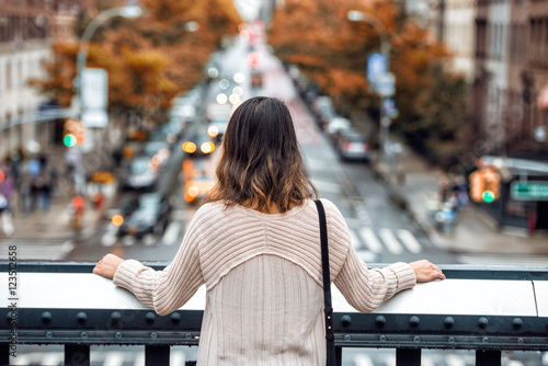 Beautiful woman travel and looking at New York City street with car traffic and yellow trees at autumn time from high point. View from the back.