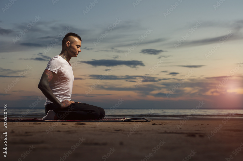Young tattoo man doing yoga exercises on the beach