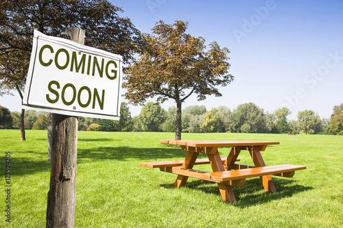 Coming soon written on a signboard near a picnic table on a green field