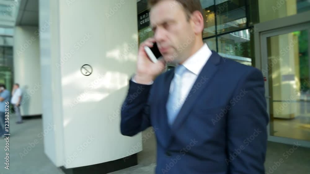 Businessman is leaving an office building and talking on the phone. Steadicam shot. Businessman is leaving an office building and talking on the phone. Steadicam shot.