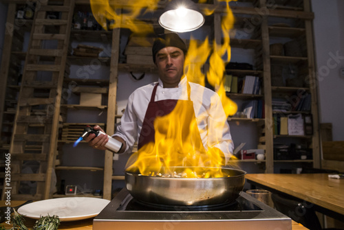 Man flambeing beef cheeks with sautéed vegetables in a pan using a kitchen torch