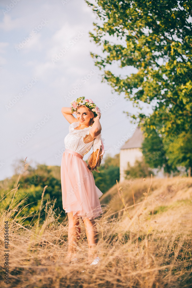 Beautiful model, slim blonde has a rest on a background of sunrise on the river near the tree and nature. White and pink dress, wreath of flowers. Air baths. Healthy lifestyle concept. Beautiful model, slim blonde has a rest on a background of sunrise on the river near the tree and nature. White and pink dress, wreath of flowers. Air baths. Healthy lifestyle concept.