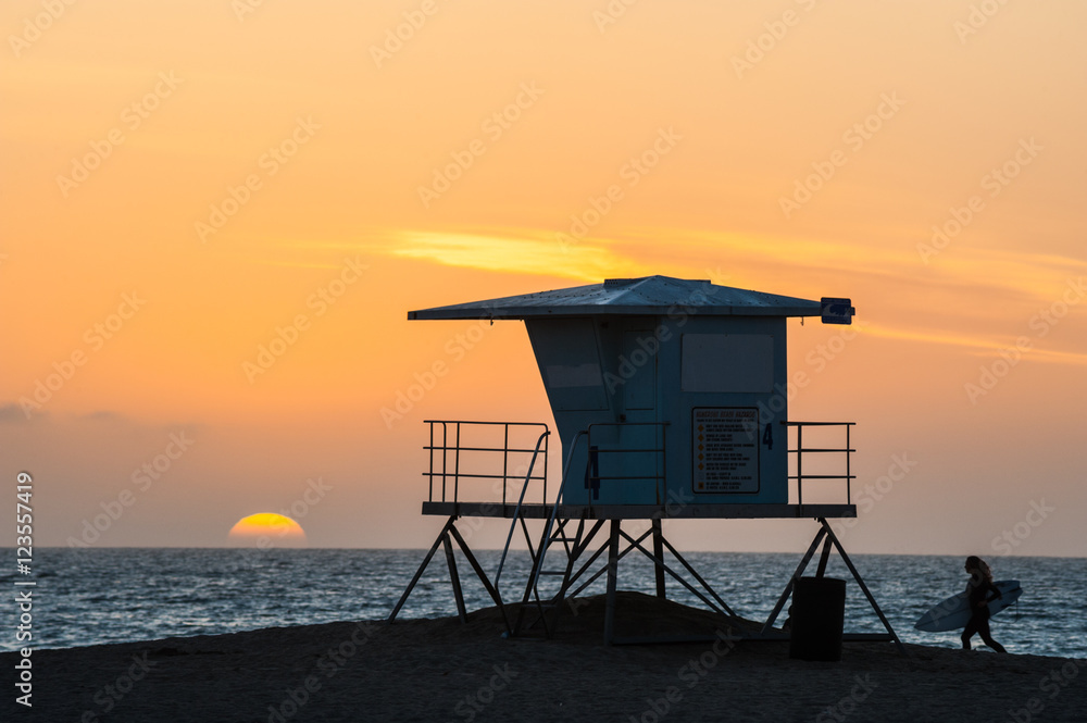 Naklejka premium Sylwetka Surfer przebiegający obok wieży ratownika podczas zachodu słońca na plaży Huntington w południowej Kalifornii