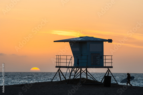 Silhouette of Surfer running past lifeguard tower during sunset on Huntington beach in southern California