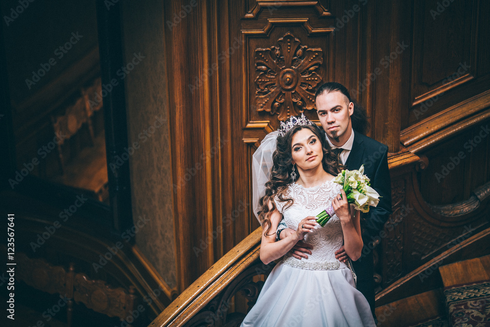 elegant gorgeous bride and stylish groom standing on wooden stairs in amazing old rich room. unusual wedding couple in retro style. luxury wedding concept. Man with long hair and piercing