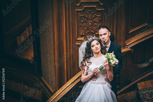 Obraz na płótnie elegant gorgeous bride and stylish groom standing on wooden stairs in amazing old rich room. unusual wedding couple in retro style. luxury wedding concept. Man with long hair and piercing w salonie