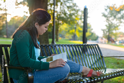 College aged girl sitting on a bench in a park reading a book
