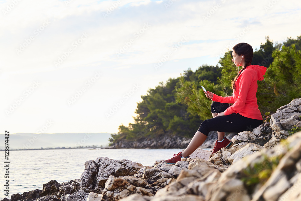 Active young woman listening to music. Runner resting from routine exercise. Happy jogger relaxing at the beach after run, sitting on the stones and looking at the sea.