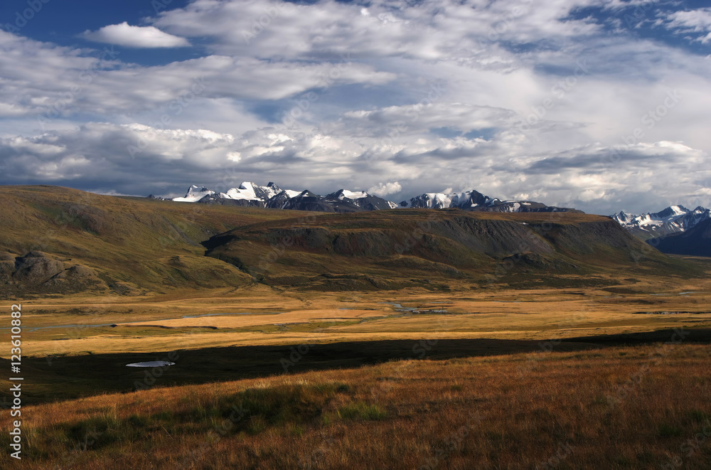 A highland river valley with yellow grass on a background of snow covered mountains and glaciers under clouds and blue sky, Plateau Ukok, Altai mountains, Siberia, Russia