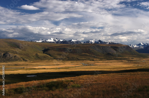 A highland river valley with yellow grass on a background of snow covered mountains and glaciers under clouds and blue sky, Plateau Ukok, Altai mountains, Siberia, Russia