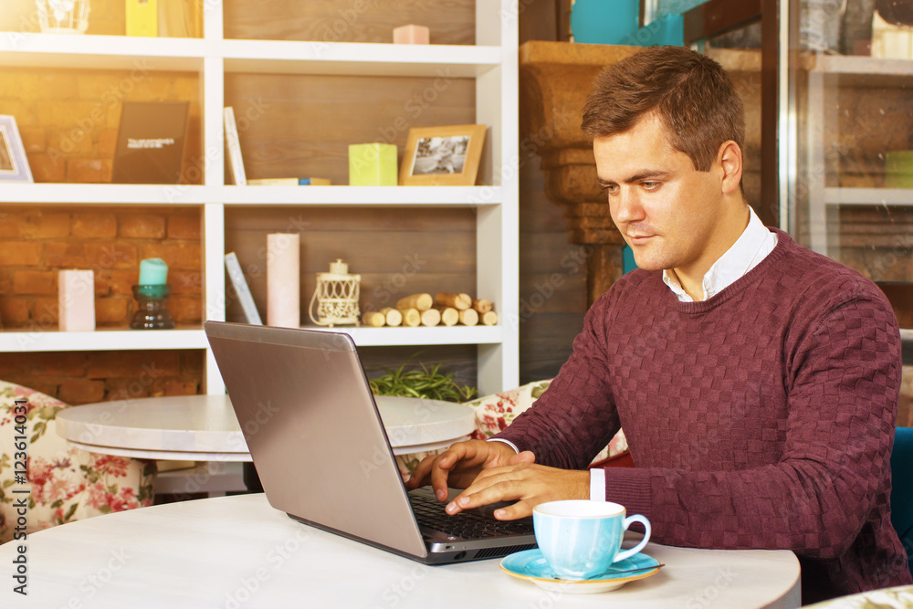 Happy young man smiling, as he works on his laptop to get all his business done early in the morning with his cup of coffee Happy young man smiling, as he works on his laptop to get all his business done early in the morning with his cup of coffee