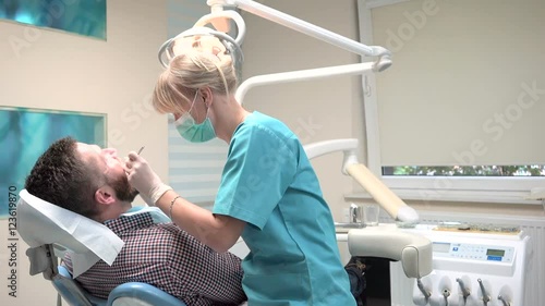 Dentist checking up teeth of patient, looks to the camera. Slider shot, left. Male patient on visit at young female dentist. Visit is in proffessional dental clinic. He is sitting on dental chair.
