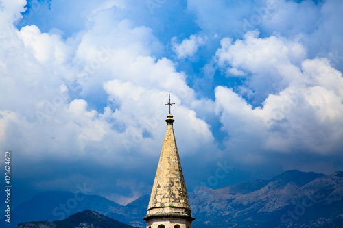 Spire of medieval catholic cathedral on background of stormy sky, dramatic clouds and mountain ranges. Saint Ivan Church in old town of Budva, Montenegro.