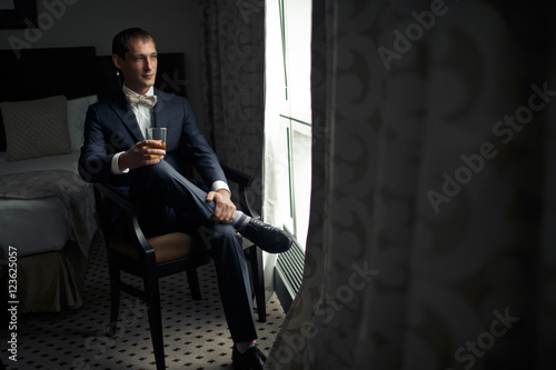 Man in dark blue suit and creamy bow tie sits in the chair behin