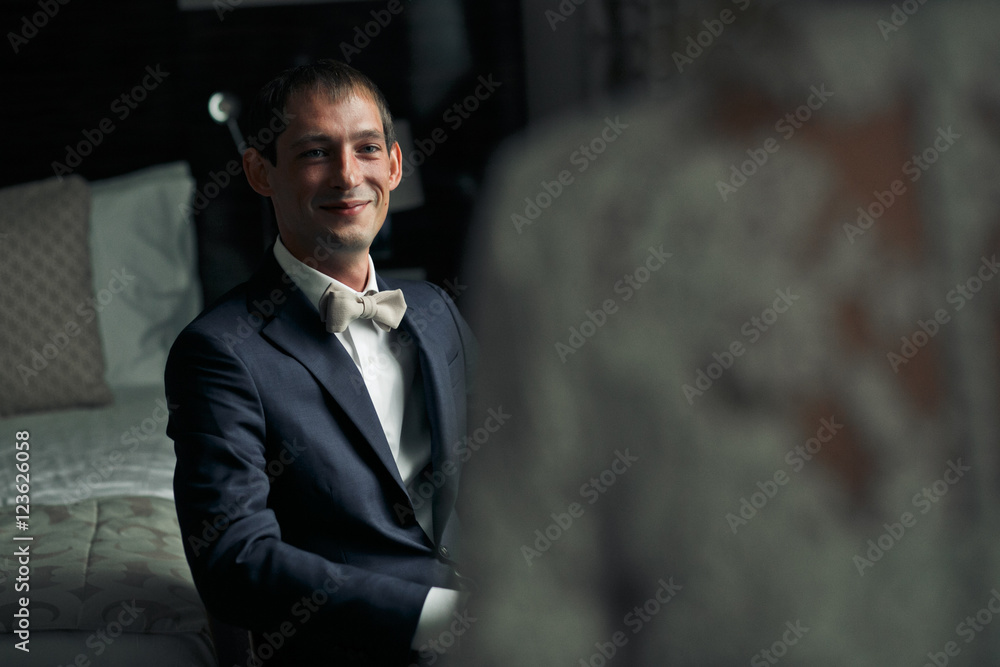 A view from bride's shoulder on a groom who smiles while sitting
