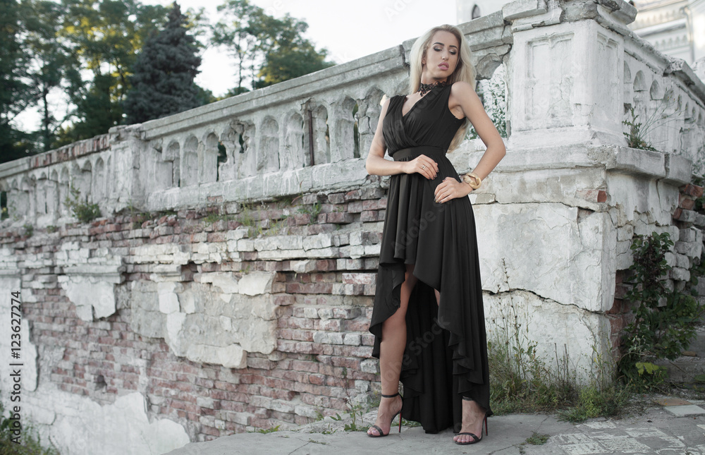 Beautiful blonde woman in luxury black dress standing by a stone wall against the staircase on a summer day