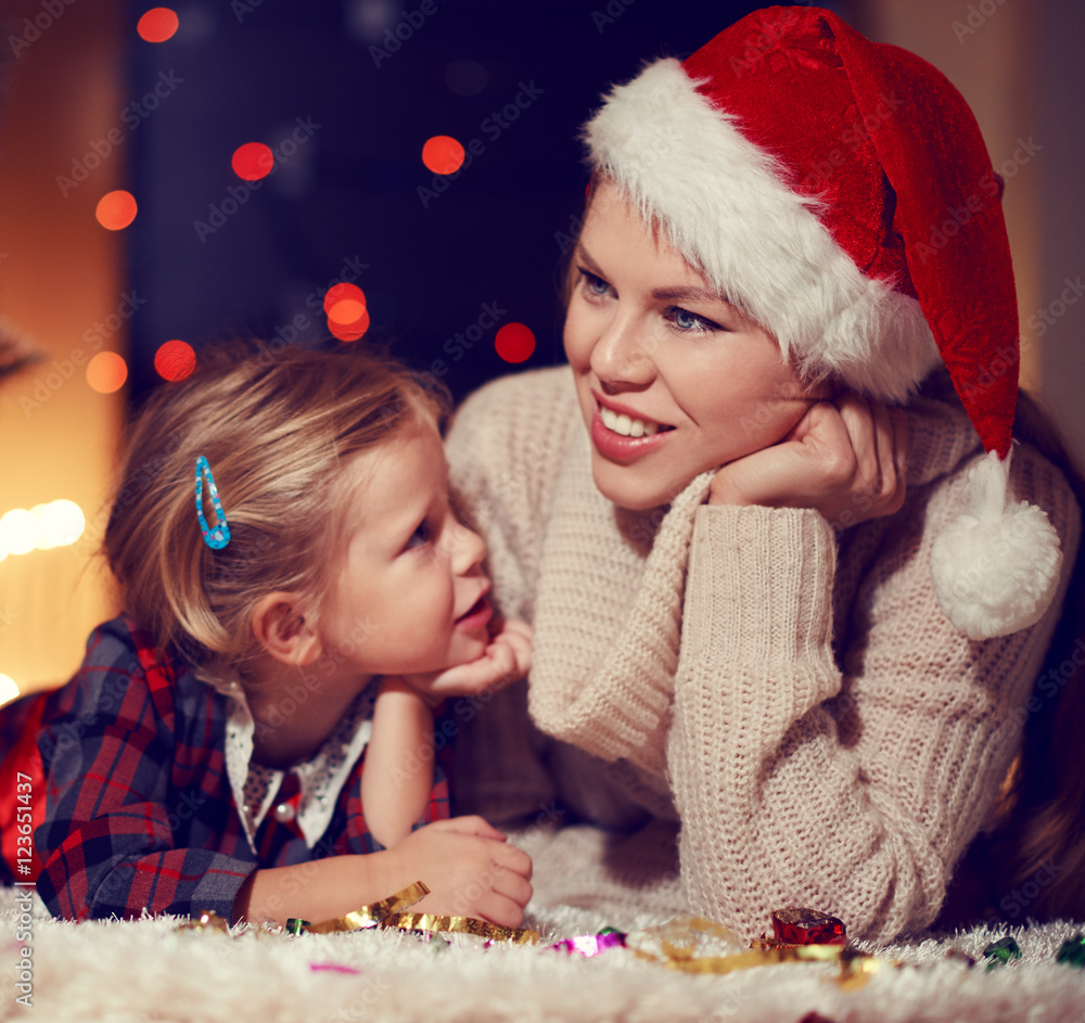 Attractive young mom in Santa hat lying on the floor with adorable child dreaming at Christmas eve. Concept of family comfort and tradition. 