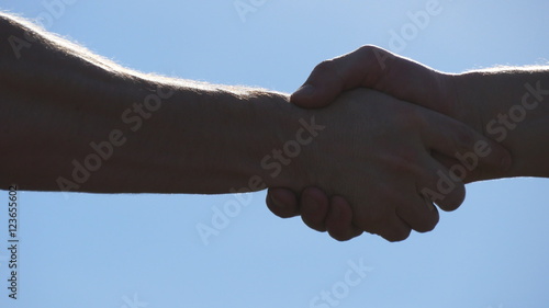 Friendly handshake of two unrecognizable muscular white men on blue sky background. Shaking of male arms outdoor. Two strong men having firm handshake outside. Teamwork and friendship. Close-up