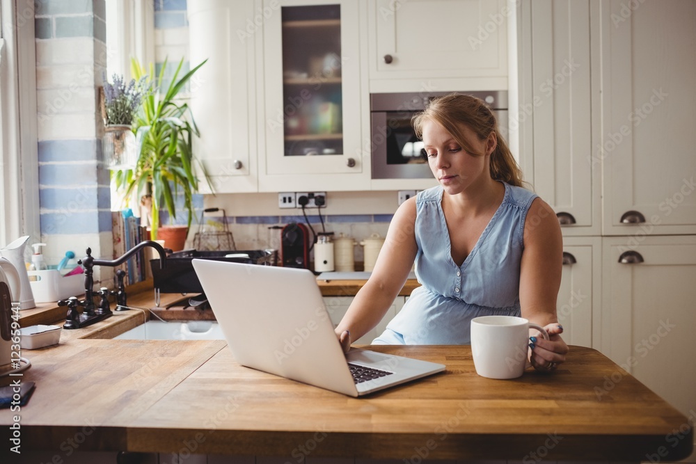 Pregnant woman using laptop while having coffee in kitchen Pregnant woman using laptop while having coffee in kitchen