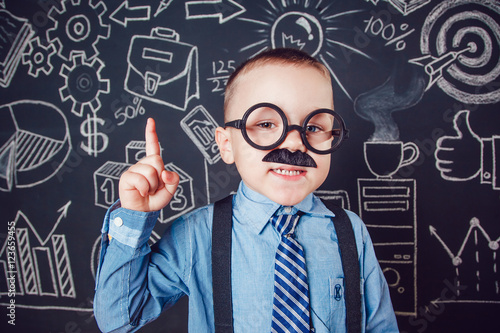 Little boy as businessman or teacher with mustache and glasses standing on dark background pattern. Wearing shirt, tie. Lifting a finger up