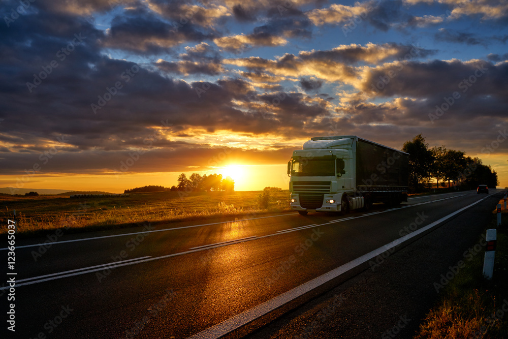 White truck driving on asphalt road along the corn field during a dramatic sunset. White truck driving on asphalt road along the corn field during a dramatic sunset.