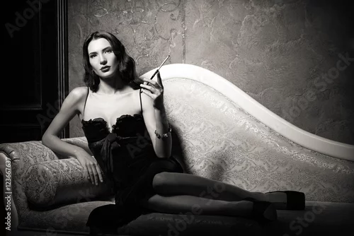 Obraz Styl filmowy noir: elegancka młoda kobieta leżąca na kanapie i paląca papierosa. Czarny i biały