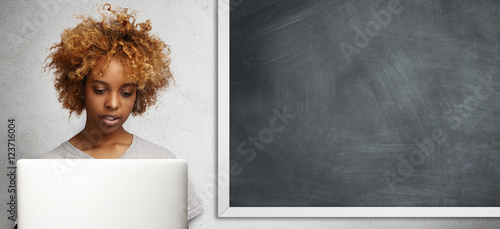 People, technology and education concept. Headshot of serious dark-skinned young teacher with curly hair using laptop computer, checking papers of her students, sitting in classroom after lessons