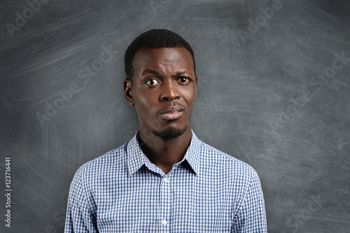 Shocked dark-skinned teacher surprised with his pupils misbehavior during his first day at school. Astonished student in shirt standing at blackboard, not ready with homework, looking displeased