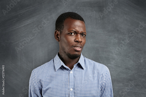 Are you serious? Irritated young dark-skinned teacher or student dressed in checkered shirt, looking dissatisfied and surprised, standing against blackboard with copy space for your advertisement