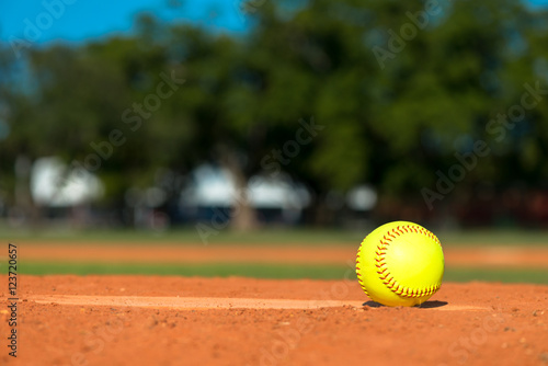 Bright fluorescent neon day-glo yellow softball on pitchers mound with red dirt field grass and trees in background