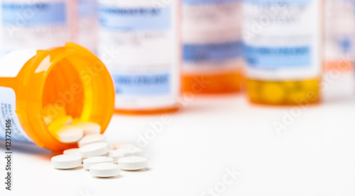 Round white prescription medication medicine pill tablets spilling from a bottle with numerous full bottles in background photo