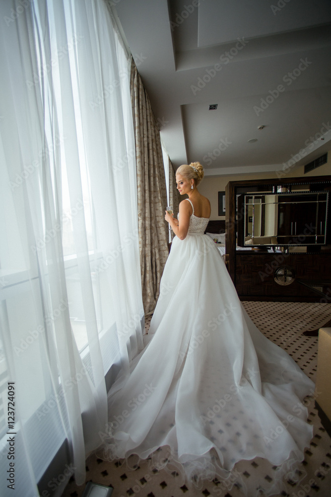 Bride stands in a hotel room with champagne flute looking throug Bride stands in a hotel room with champagne flute looking throug
