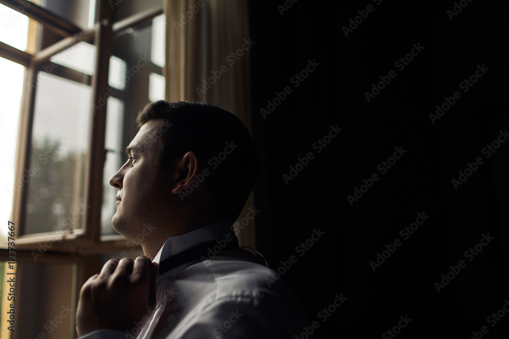 Brunette groom is standing next to the window Brunette groom is standing next to the window
