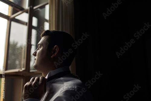 Brunette groom is standing next to the window © nastasenko