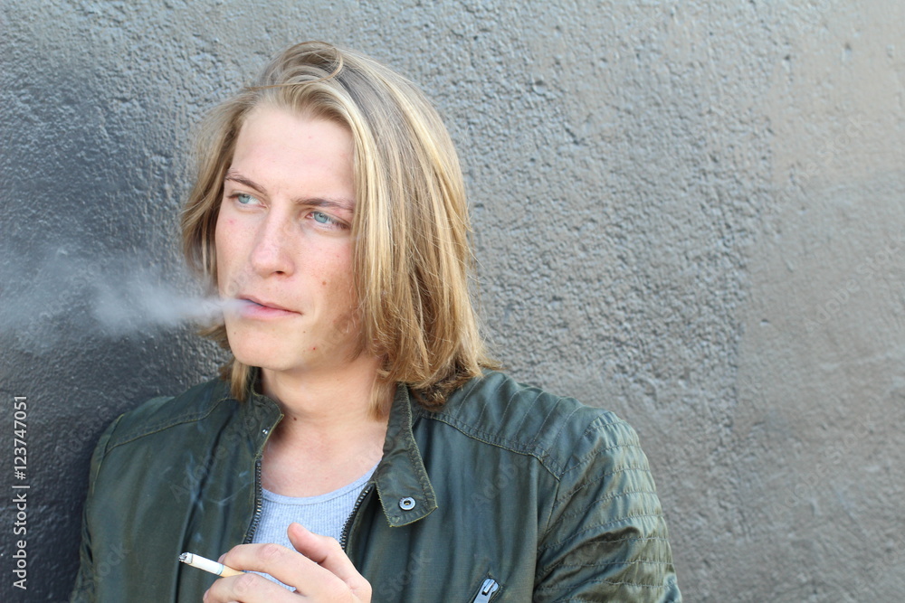 Young blond man with cigarette in mouth, isolated over gray background with copy space 