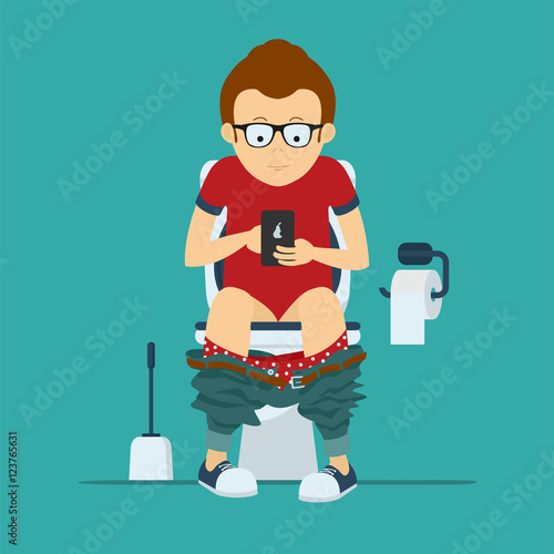 Guy hipster sits on toilet bowl with phone in hands. Toilet bowl, toilet paper and brush for toilet bowl.