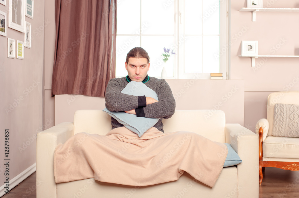 Sad and tired young man in sweater sitting on the sofa at home with pillow and plaid. Concept of illness.