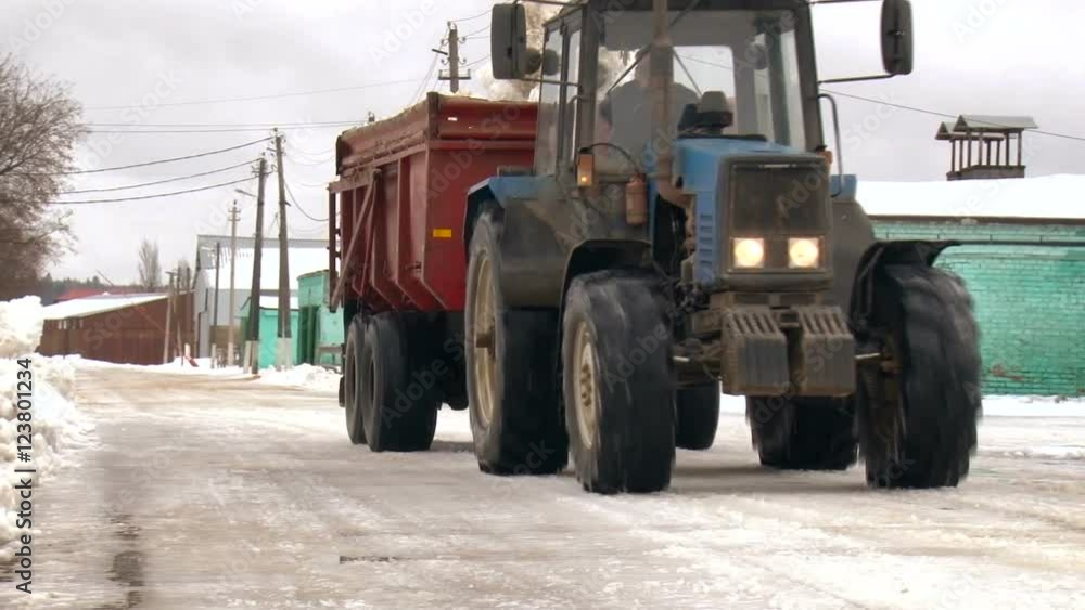 Countryside. Farming Area. Village Street. Tractor With Red Container on Trailer Moving on the Road. 1 Storey Wooden Houses. Winter Season Countryside. Farming Area. Village Street. Tractor With Red Container on Trailer Moving on the Road. 1 Storey Wooden Houses. Winter Season