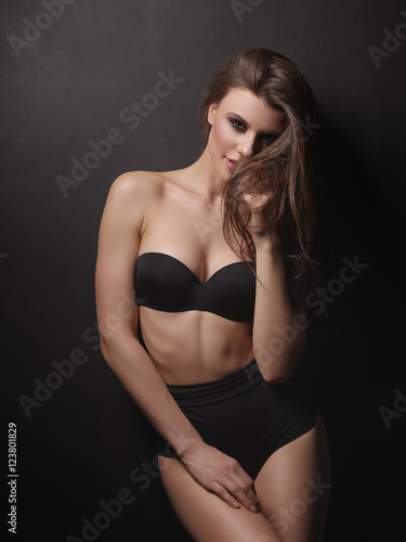 Playful and attractive girl with thick brown hair and sexy gorgeous body is posing in the black underwear on the dark background in the studio © maksimvostrikov