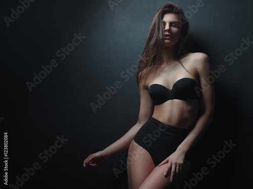 Seductive, beautiful and attractive girl with loose brown hair and sexy gorgeous figure is posing in the black seamless underwear on the dark background in the studio © maksimvostrikov